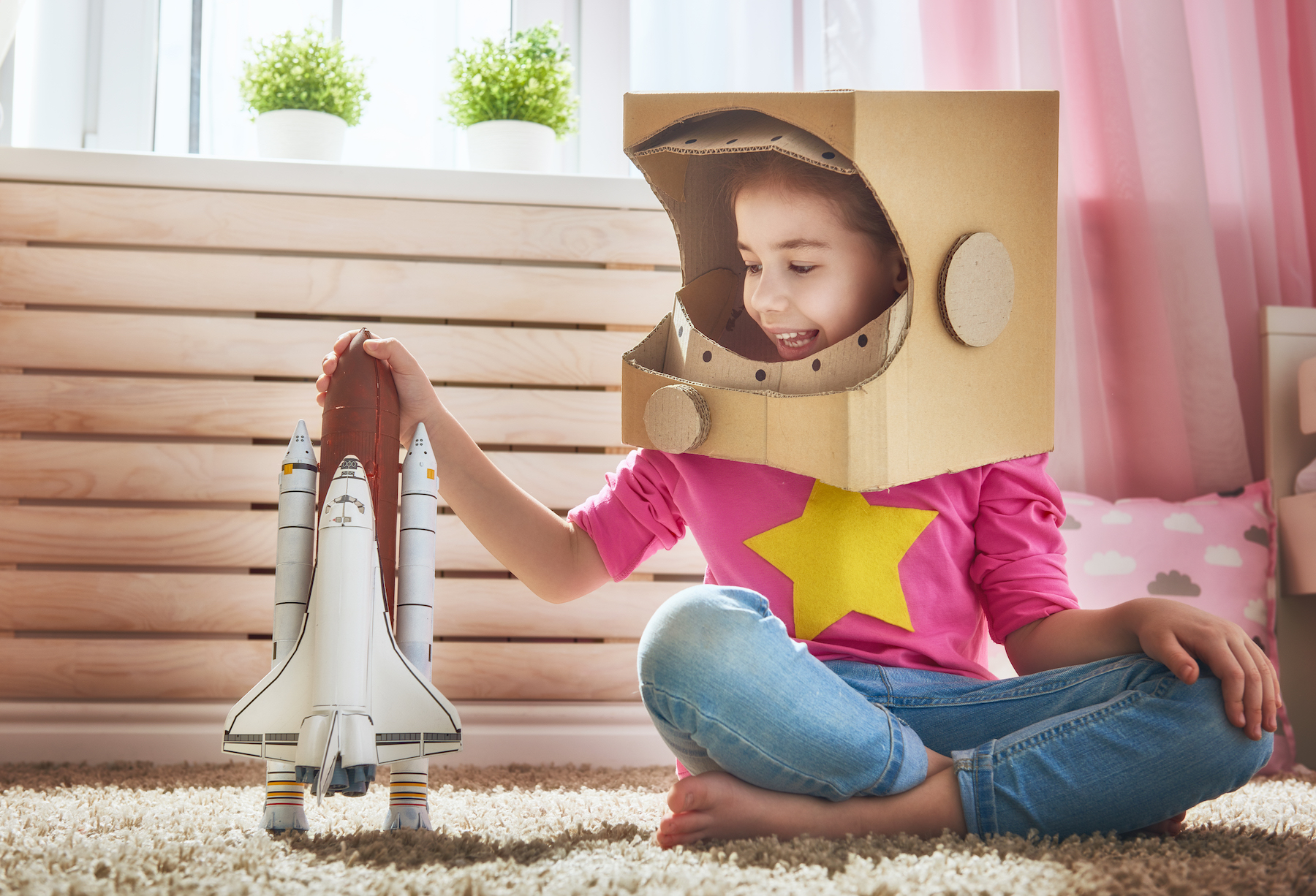 Child girl in an astronaut costume with toy rocket playing and dreaming of becoming a spacemen. Portrait of funny kid near windows.